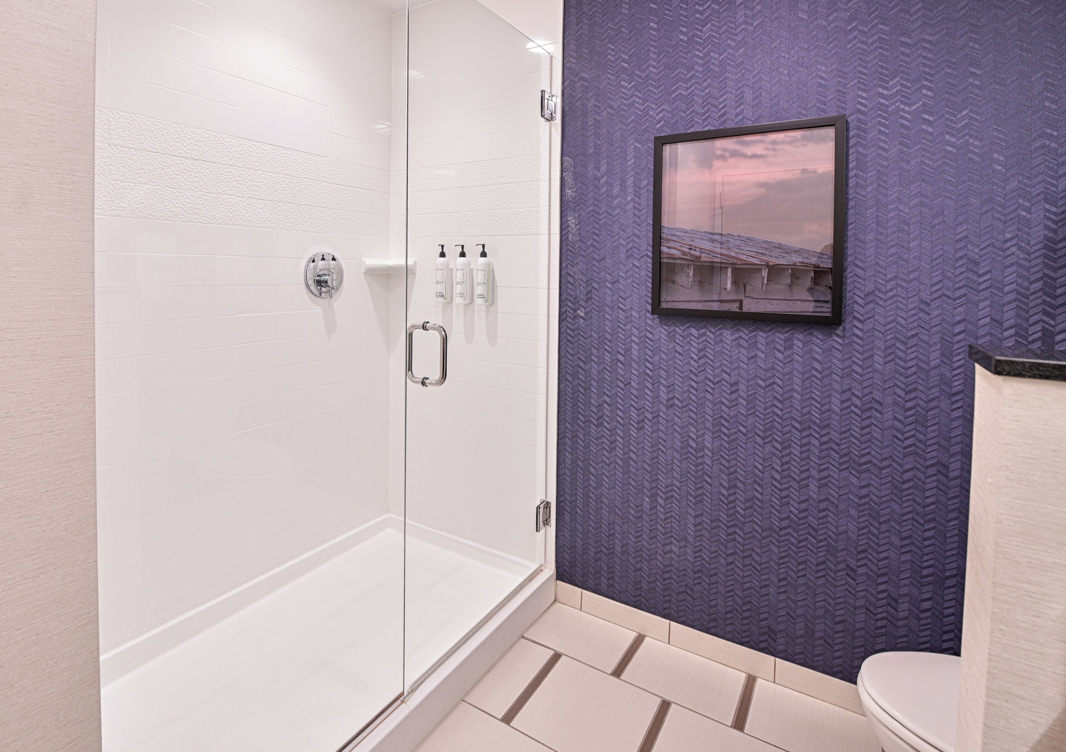 A bathroom with a glass shower enclosure, a purple textured wall, and a framed picture.