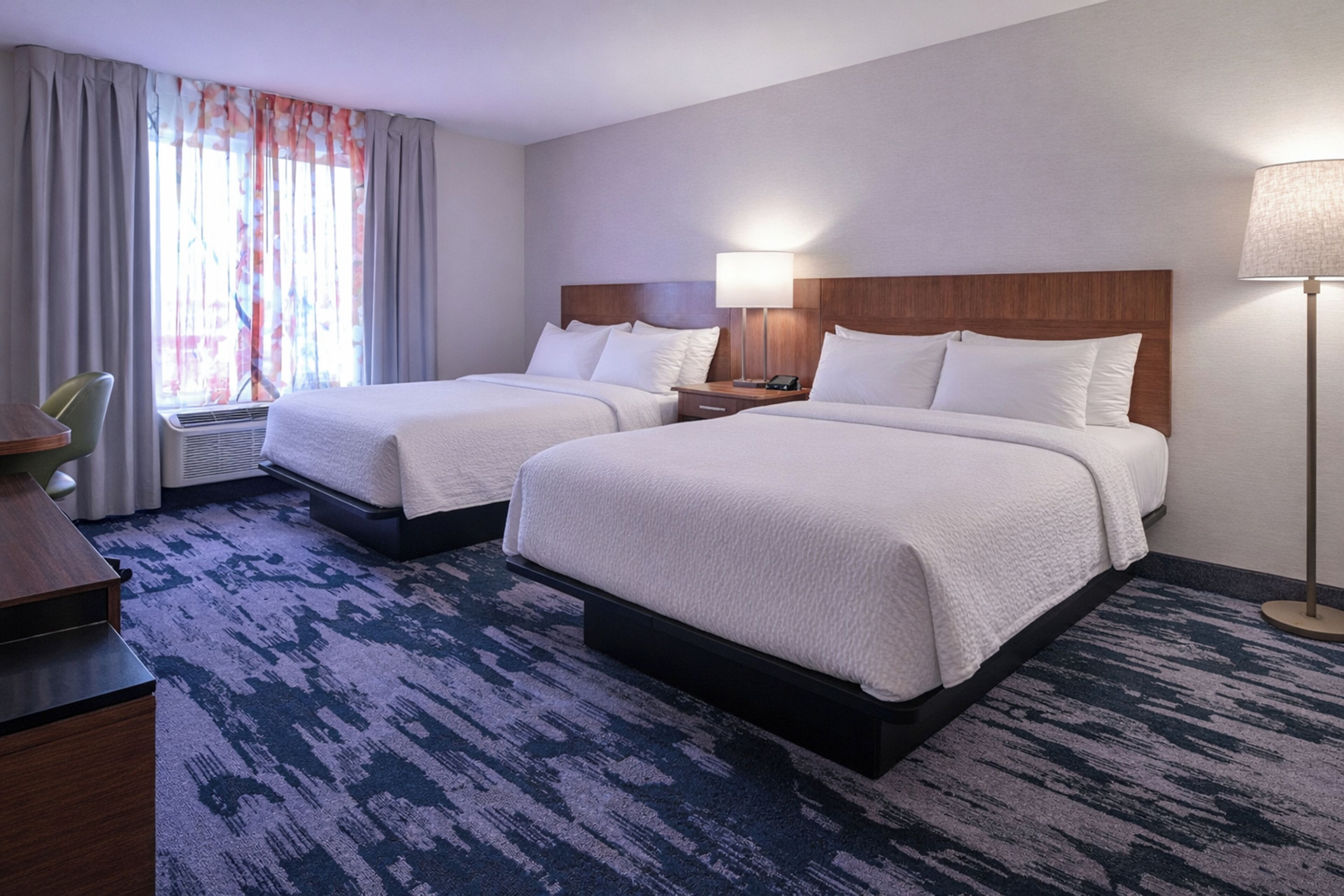 Habitación de hotel con dos camas, un escritorio, una silla, una lámpara y una ventana con cortinas.