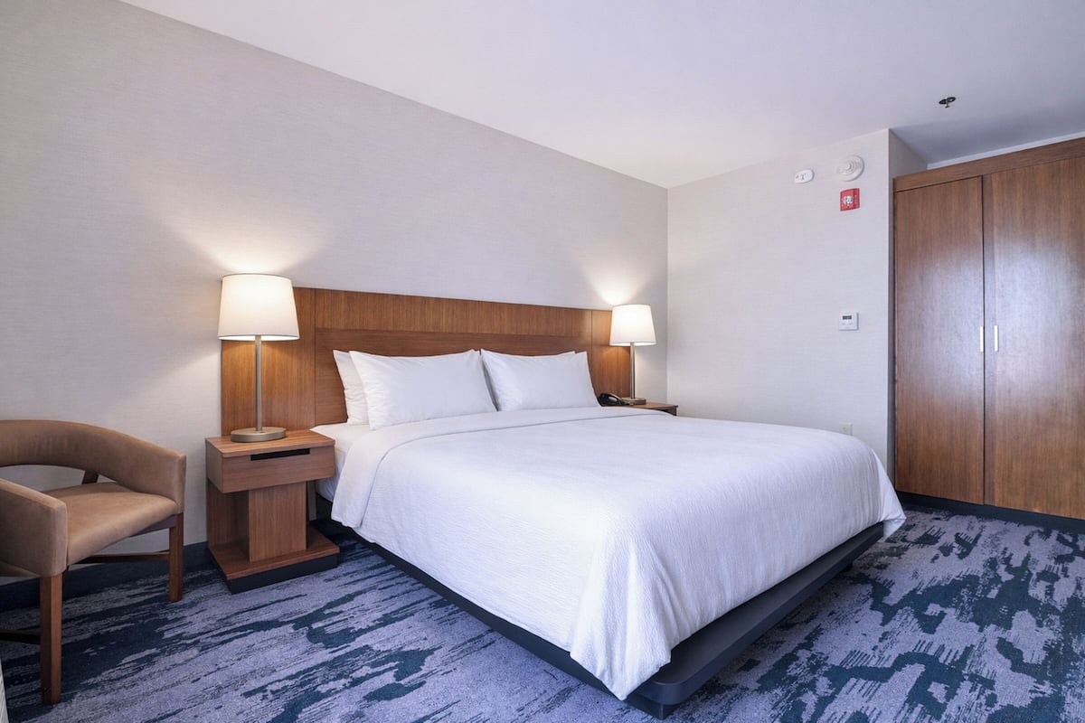 A hotel room with a bed, two bedside lamps, a chair, a wooden wardrobe, and a carpeted floor.