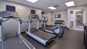 Et fitnessrum med løbebånd, et tv ophængt på væggen og et stativ med håndvægte.
