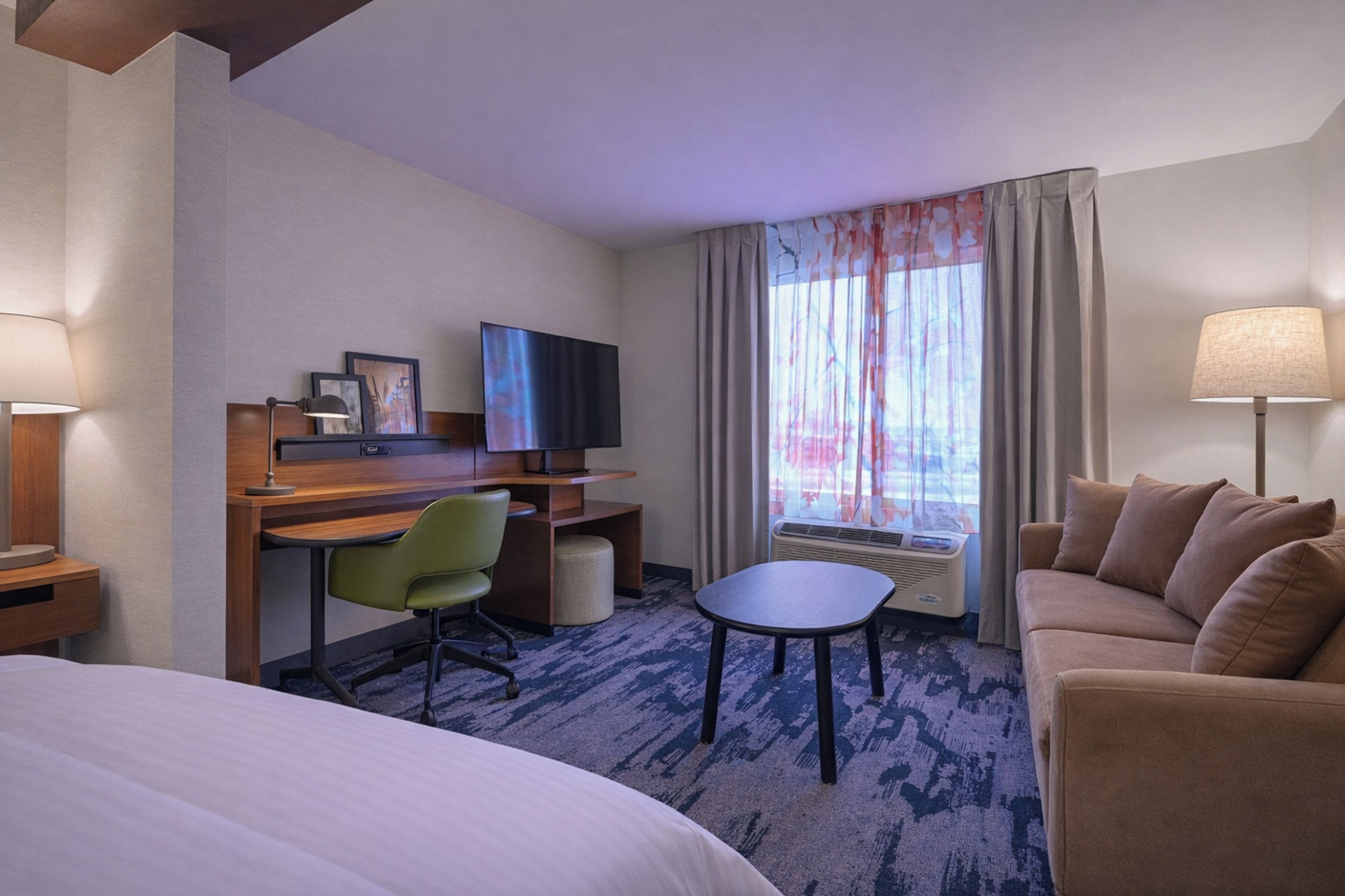 A hotel room with a bed, a desk with a chair, a TV, a sofa, and a small table.