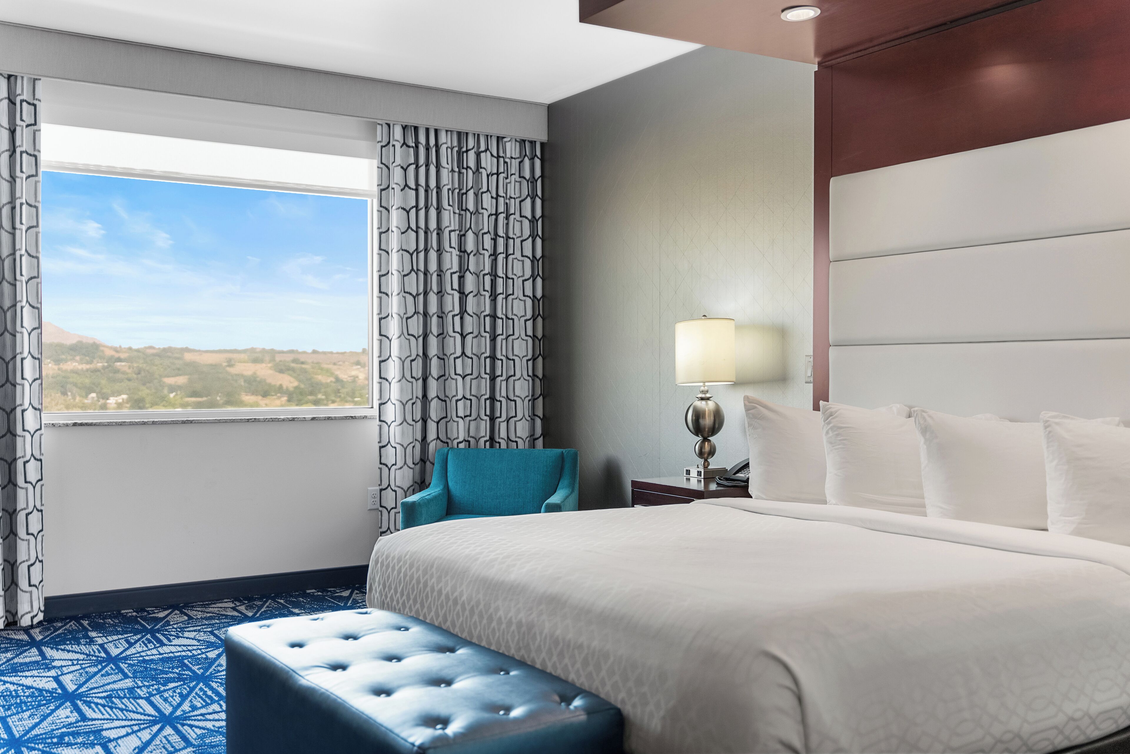 A hotel room with a large bed, a blue ottoman, a nightstand with a lamp, and a view of the outdoors through a window with patterned curtains.