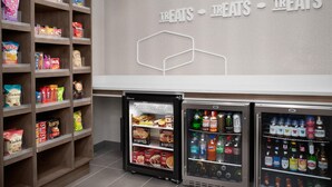 Zona de exhibición de aperitivos y bebidas, con estantes llenos de opciones y dos dispensadores de bebidas.