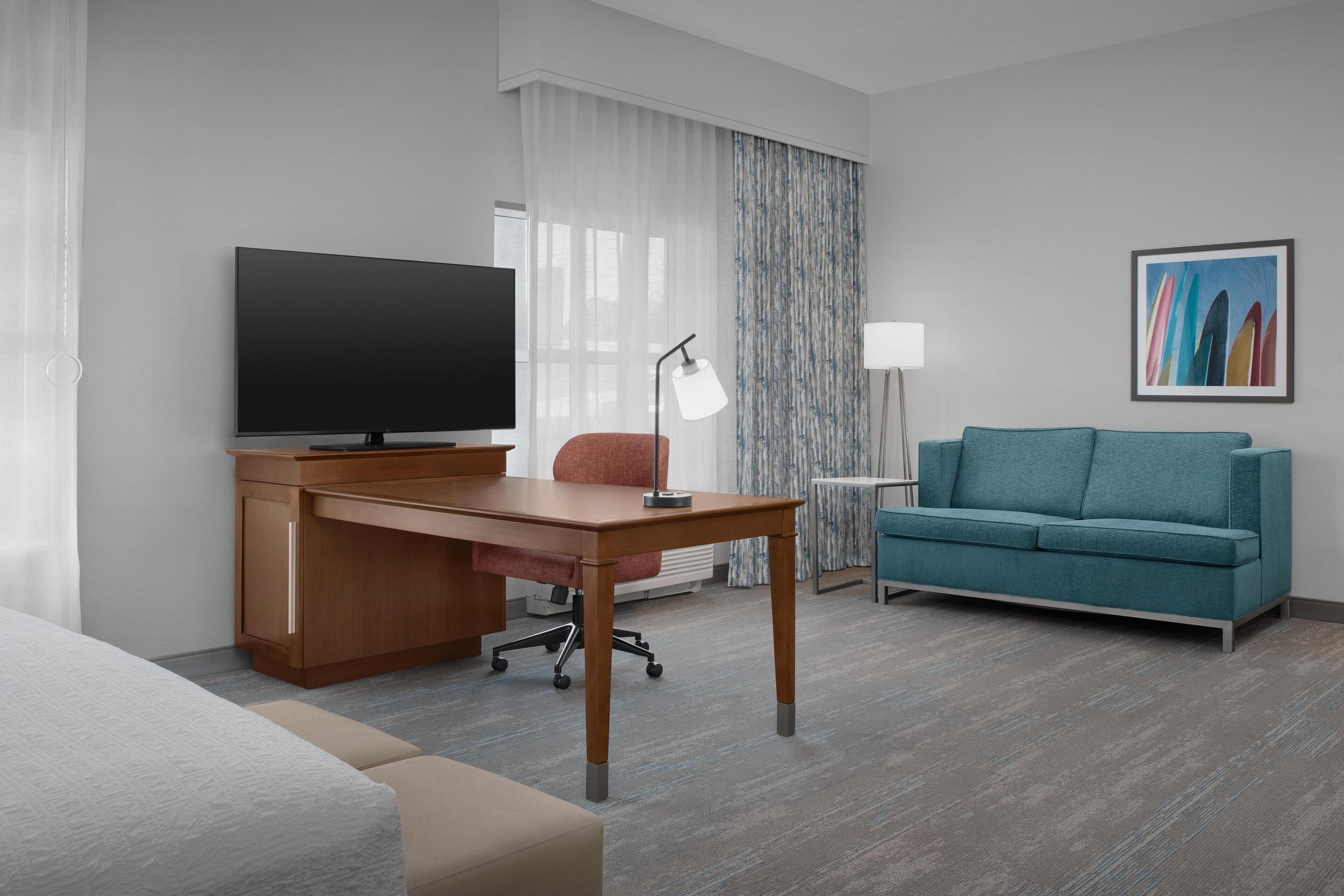 A hotel room with a bed, a desk with a TV, a teal sofa, and a painting on the wall.