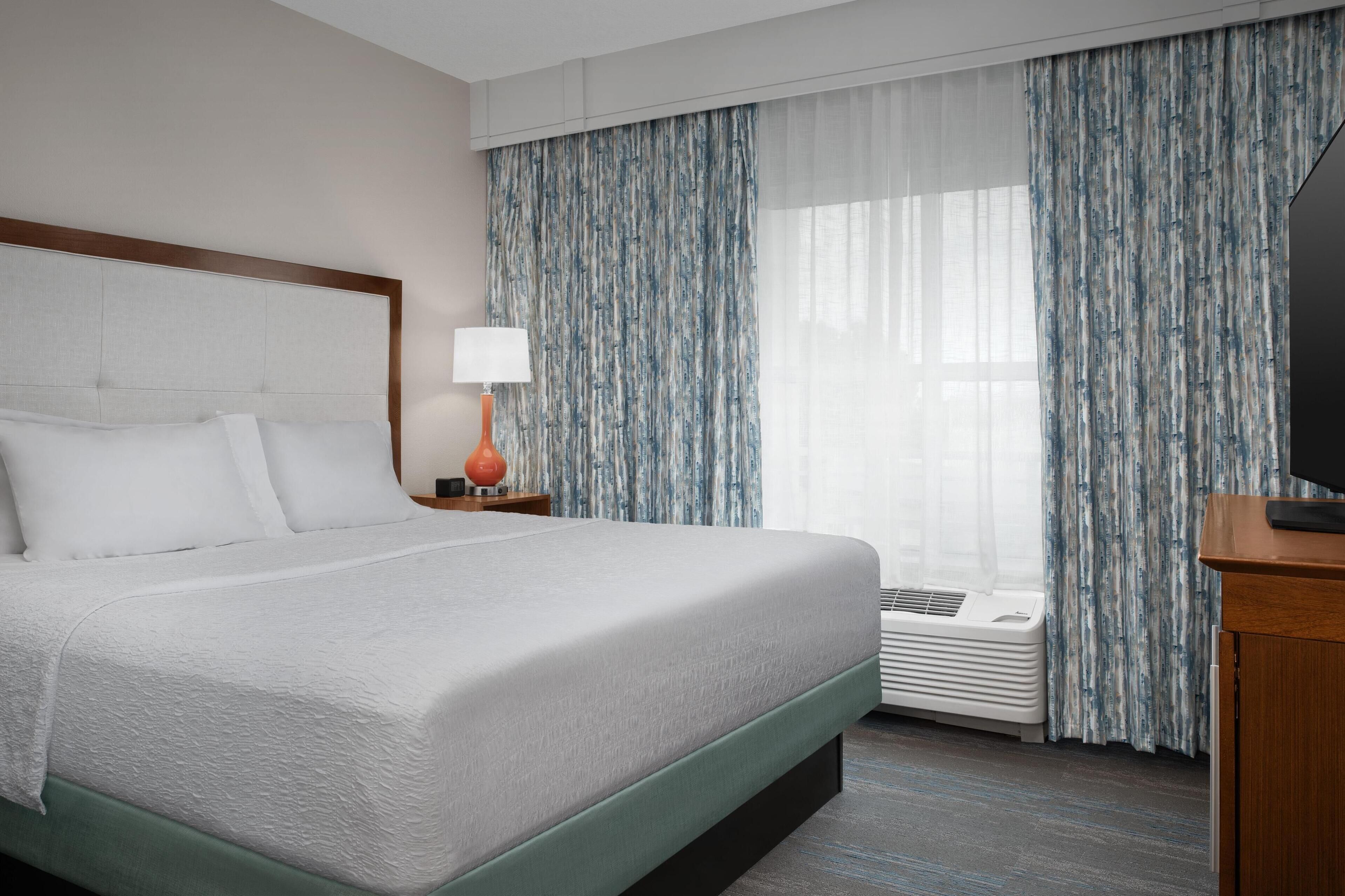 Quarto de hotel com uma cama grande, mesa de cabeceira com abajur, televisão e janela com cortinas.