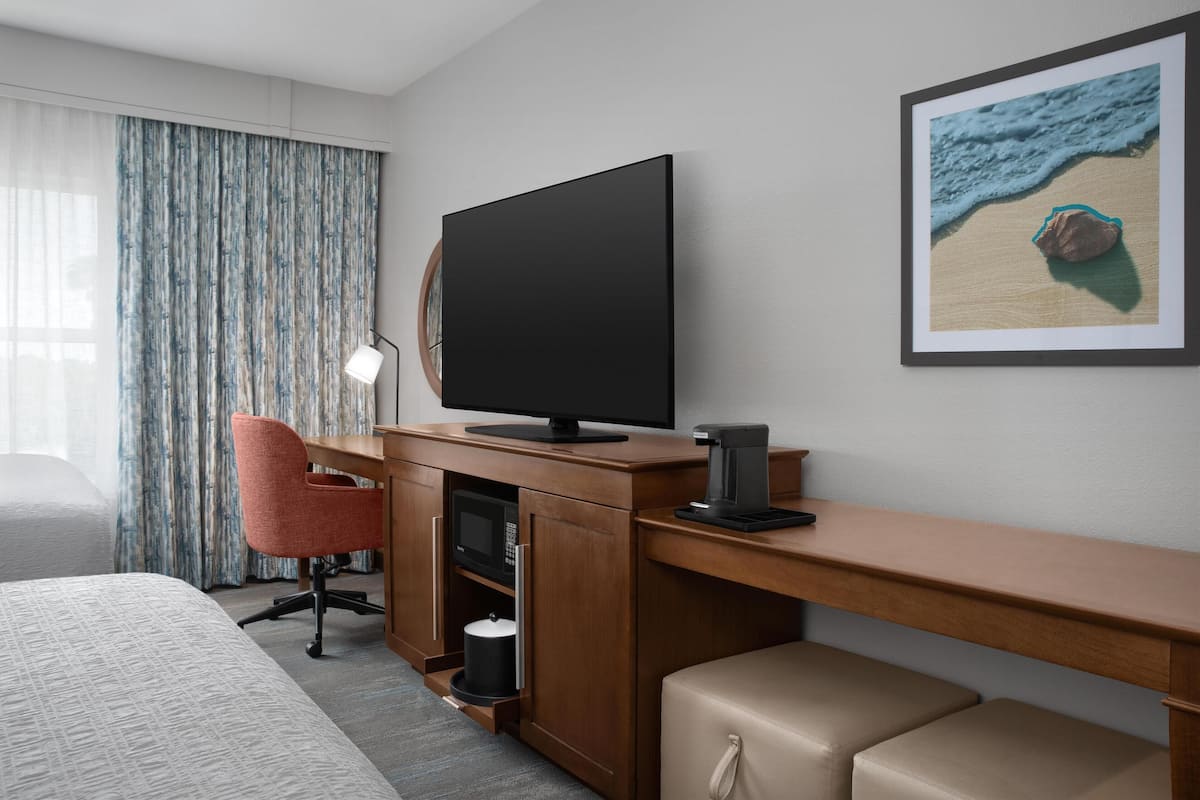 Ein Hotelzimmer mit einem Bett, einem Schreibtisch mit Fernseher, einer Kaffeemaschine, einem Sessel und einem Bild von einem Strand.