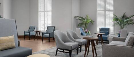 A spacious living area with a grey sofa, two armchairs, a round table, and a coffee table. There are two large windows with blinds, and potted plants are placed near the windows.