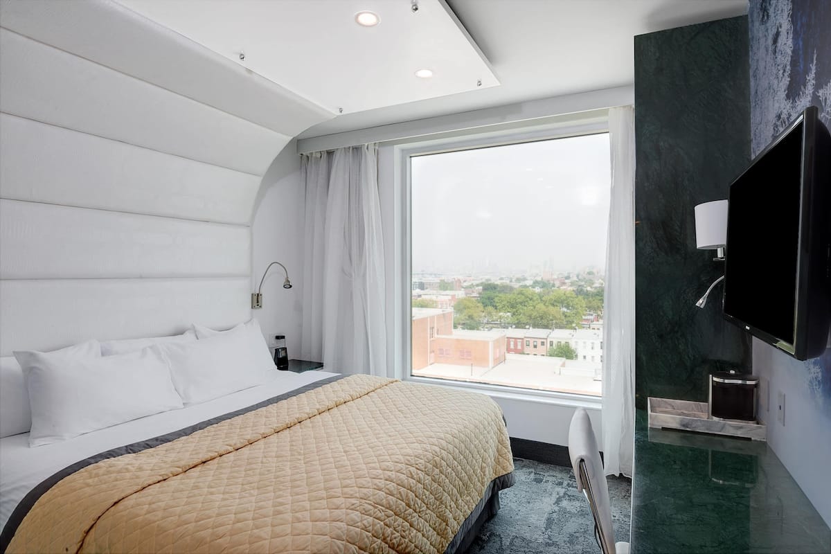 A hotel room with a large bed, a window with a city view, a bathroom with a mirror and sink, and a TV mounted on the wall.