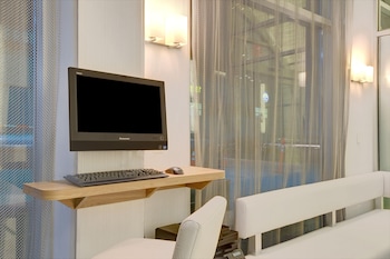 A hotel room with a bed, a desk with a laptop, a window with a view of buildings, and a nightstand with a lamp and a vase of flowers. at Wyndham Garden Brooklyn Sunset Park
