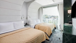 Habitación de hotel con dos camas, un ventanal grande, una lámpara en el techo, un televisor y un baño con espejo y lavamanos.