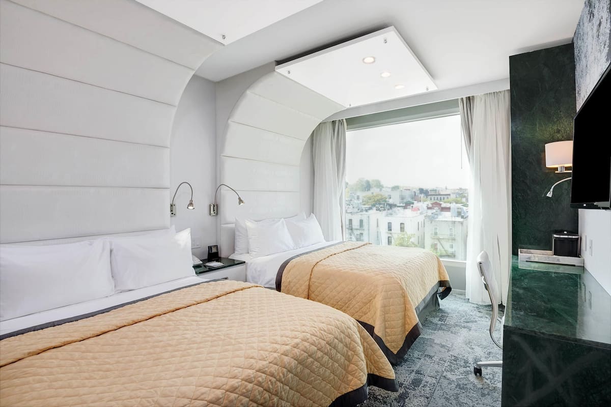 A hotel room with two beds, a large window, a ceiling light, a TV, and a bathroom with a mirror and sink.