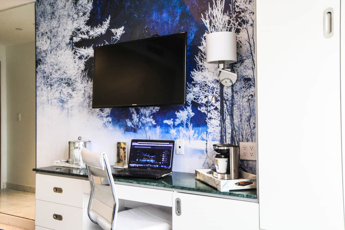 A modern office space with a flat-screen TV mounted on a wall with a winter forest mural, a laptop on a desk, a white chair, and a coffee machine.