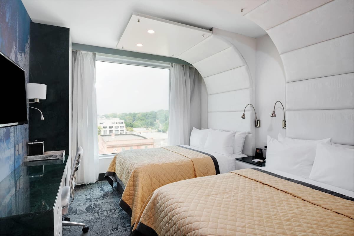 A hotel room with two beds, a TV, a desk, and a large window.