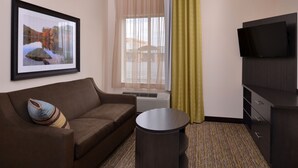 A hotel room with a brown sofa, a round side table, a flat-screen TV, a framed picture on the wall, and a window with sheer curtains. - Candlewood Suites MORGANTOWN-UNIV WEST VIRGINIA by IHG (Morgantown)