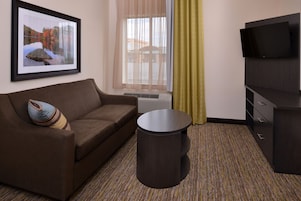 Habitación de hotel con un sofá marrón, una mesita redonda, un televisor de pantalla plana, un cuadro enmarcado en la pared y una ventana con cortinas de gasa.