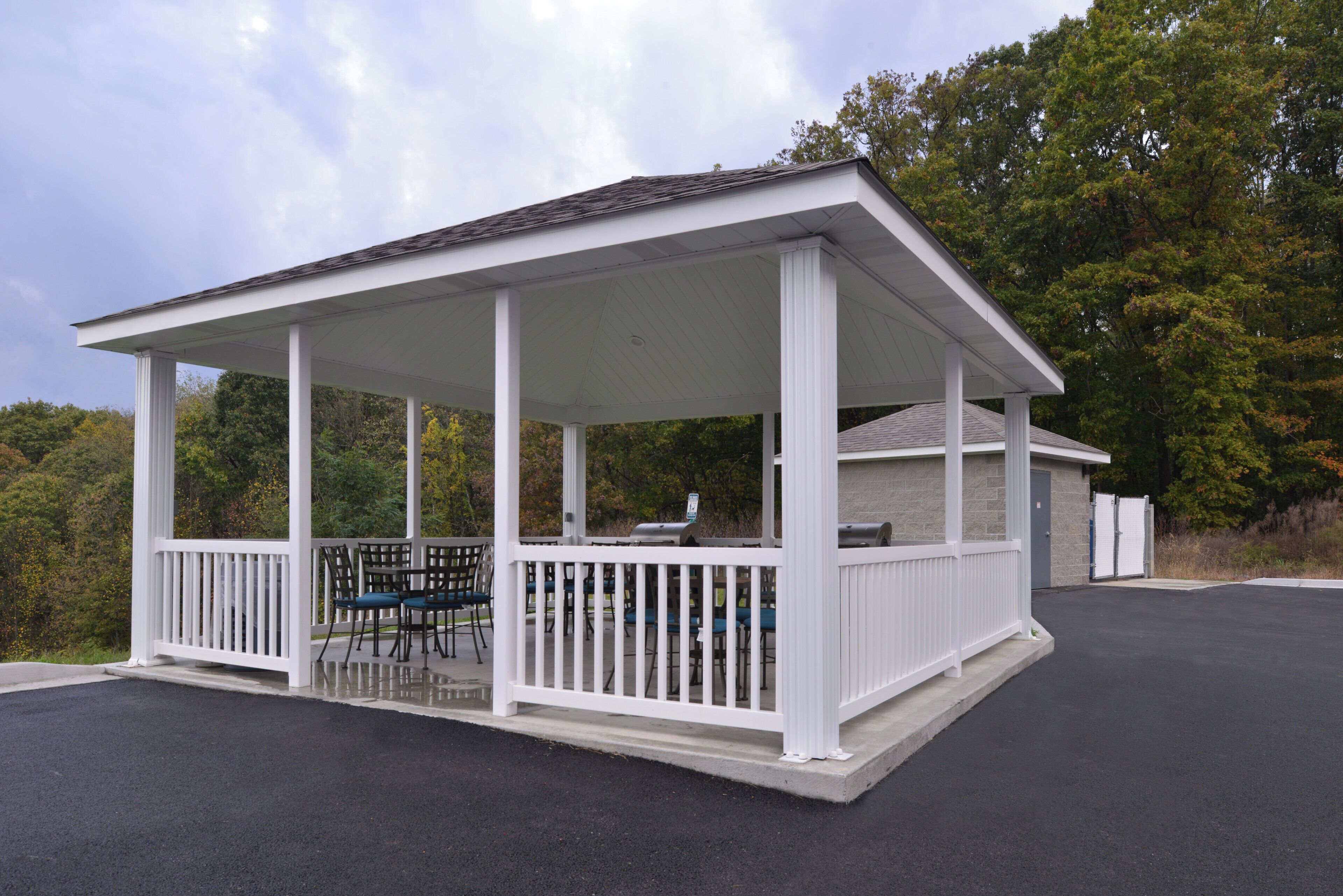 A covered outdoor seating area with a grill, surrounded by white railings and trees in the background.