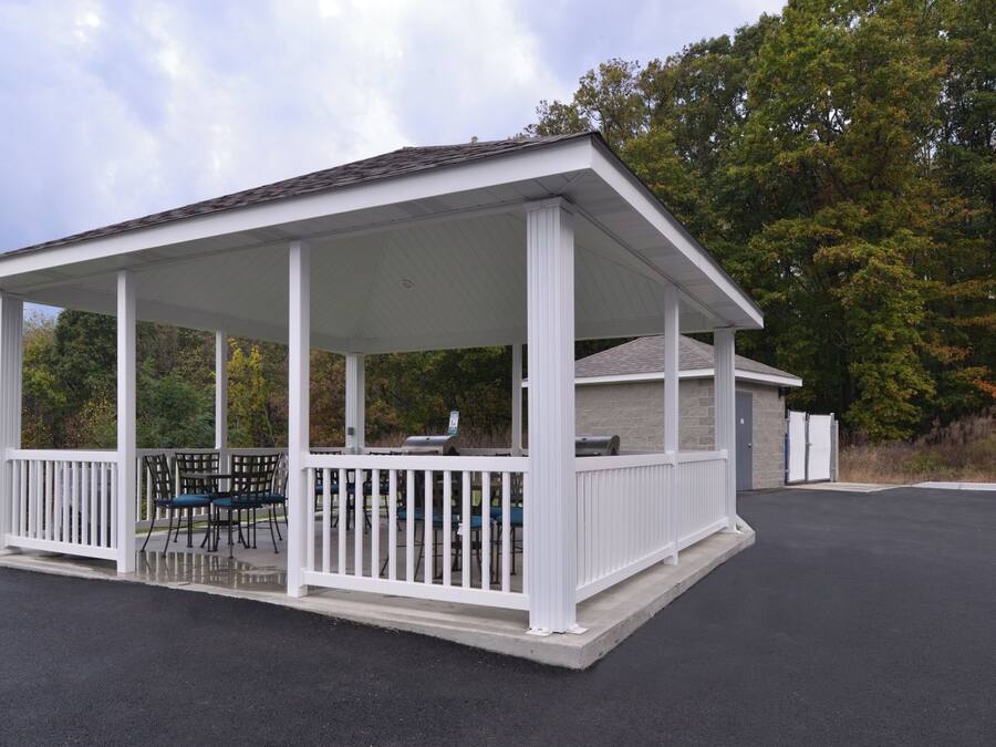 A covered outdoor seating area with a grill, surrounded by white railings and trees in the background.