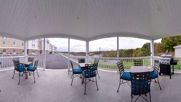 A covered outdoor patio with tables and chairs, a barbecue grill, and a view of a residential area.