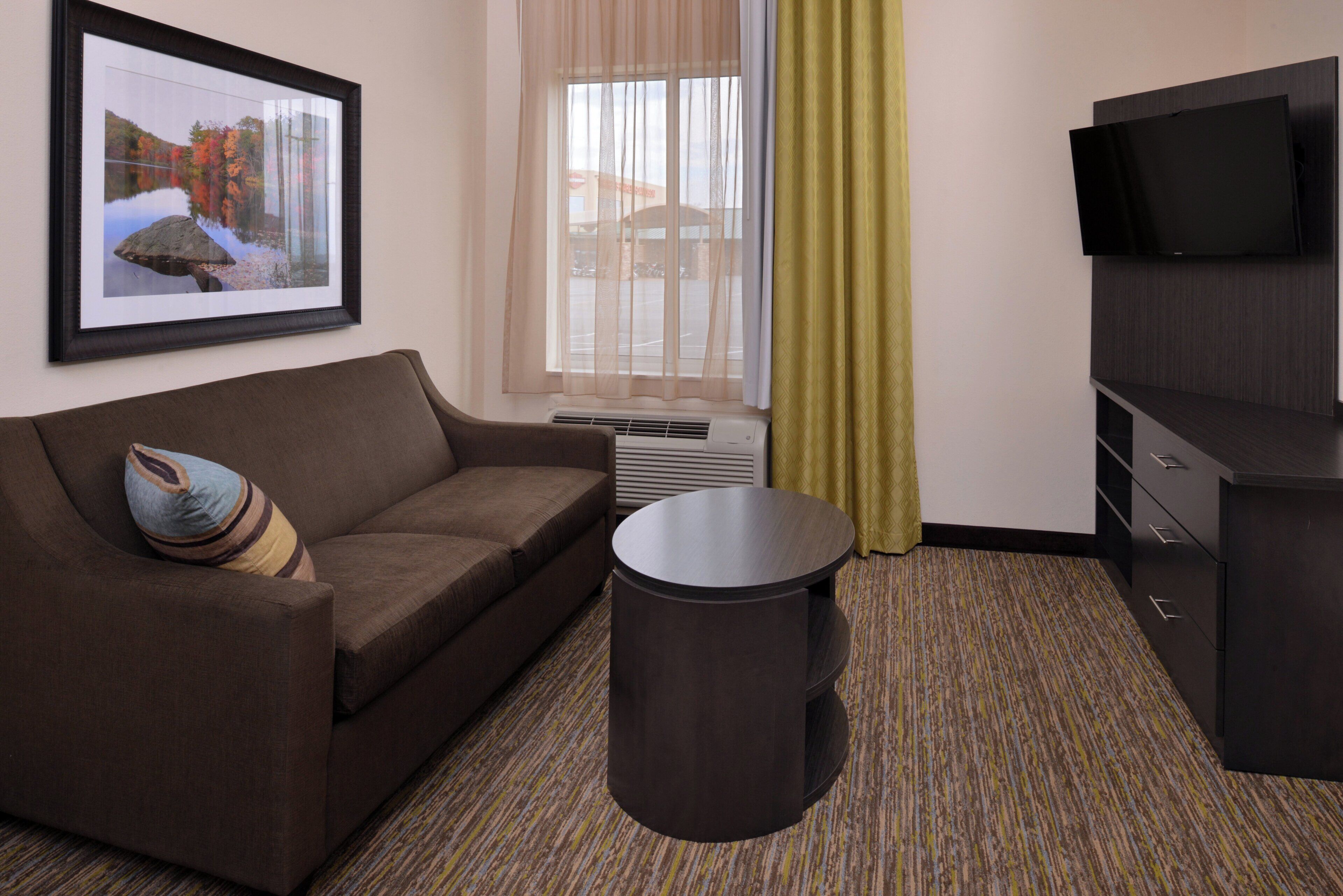 A hotel room with a brown sofa, a small round table, a TV on a cabinet, a framed picture on the wall, and a window with sheer curtains.