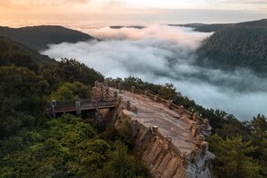 Uma ponte de pedra com grades que leva até a beira de um penhasco, com vista para um vale coberto por densas florestas e uma camada de nuvens abaixo.