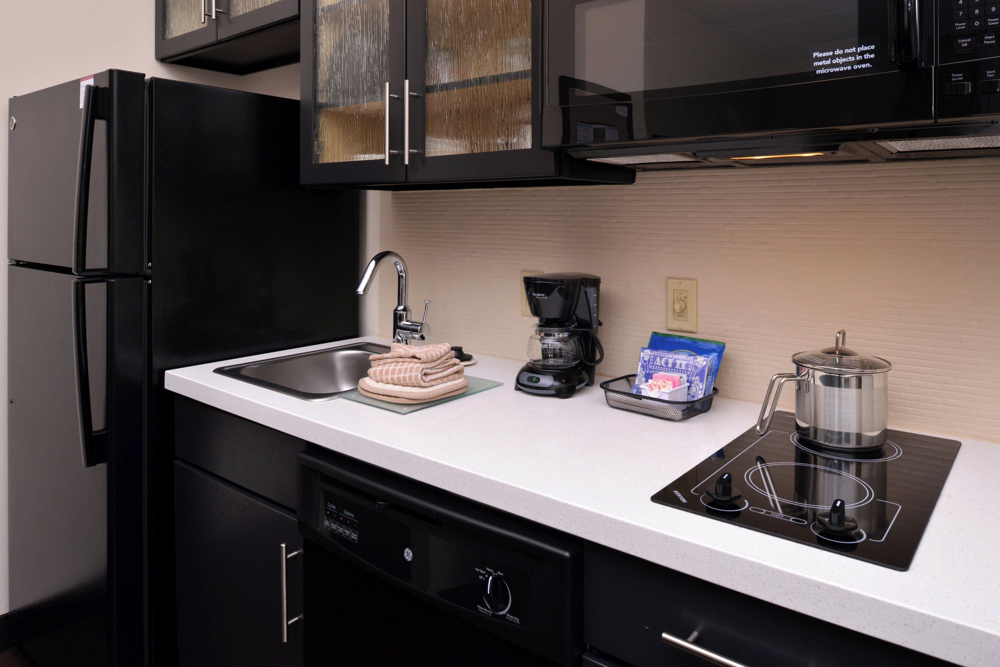A kitchen with a black refrigerator, a microwave, a coffee maker, and a kettle on an induction cooktop.