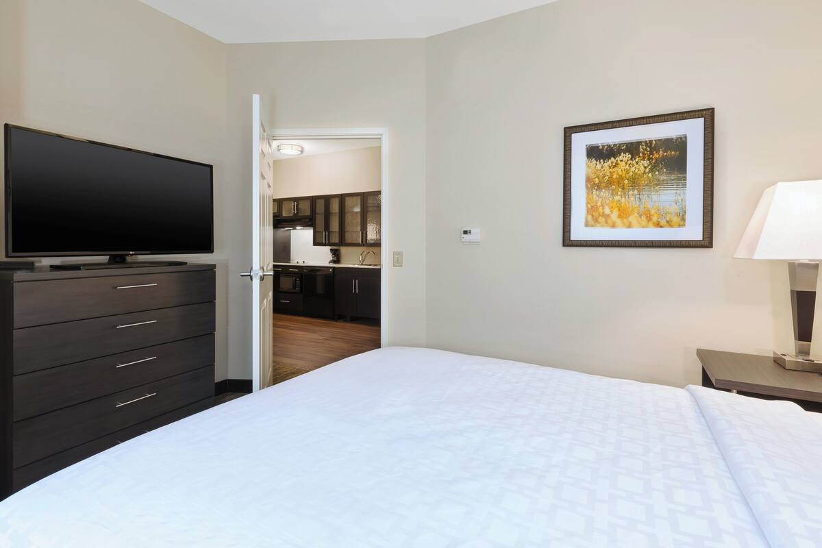 Ein Hotelzimmer mit einem großen Bett, einem Fernseher auf einer Kommode, einem gerahmten Bild an der Wand und Blick zur Küche.