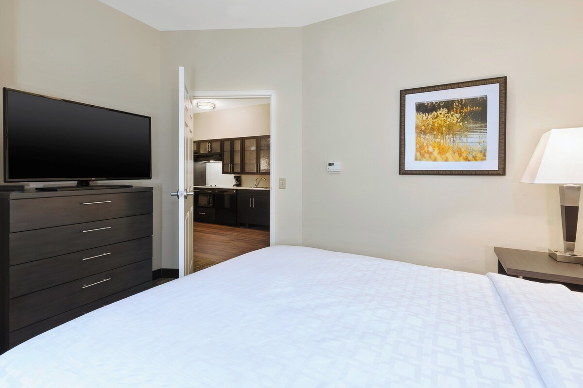 Quarto de hotel com uma cama grande, uma televisão em uma cômoda, um quadro na parede e vista para a cozinha.