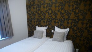 A double bed with white bedding and two pillows, a window with curtains, and a patterned wallpaper on the wall. - White Princess (Koksijde)