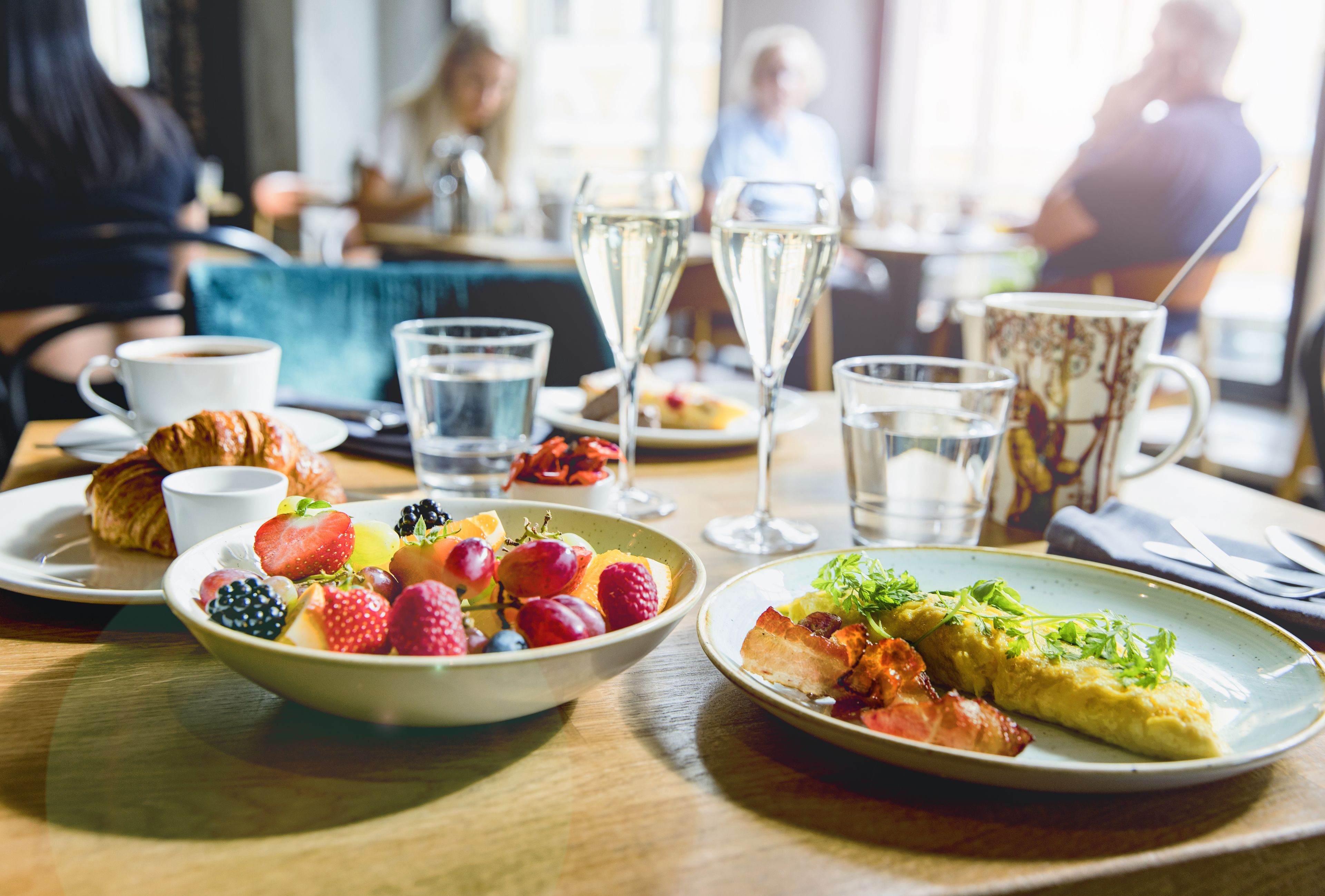 A dining table with a croissant, fruit bowl, and plates of food, glasses of water, and a coffee cup.