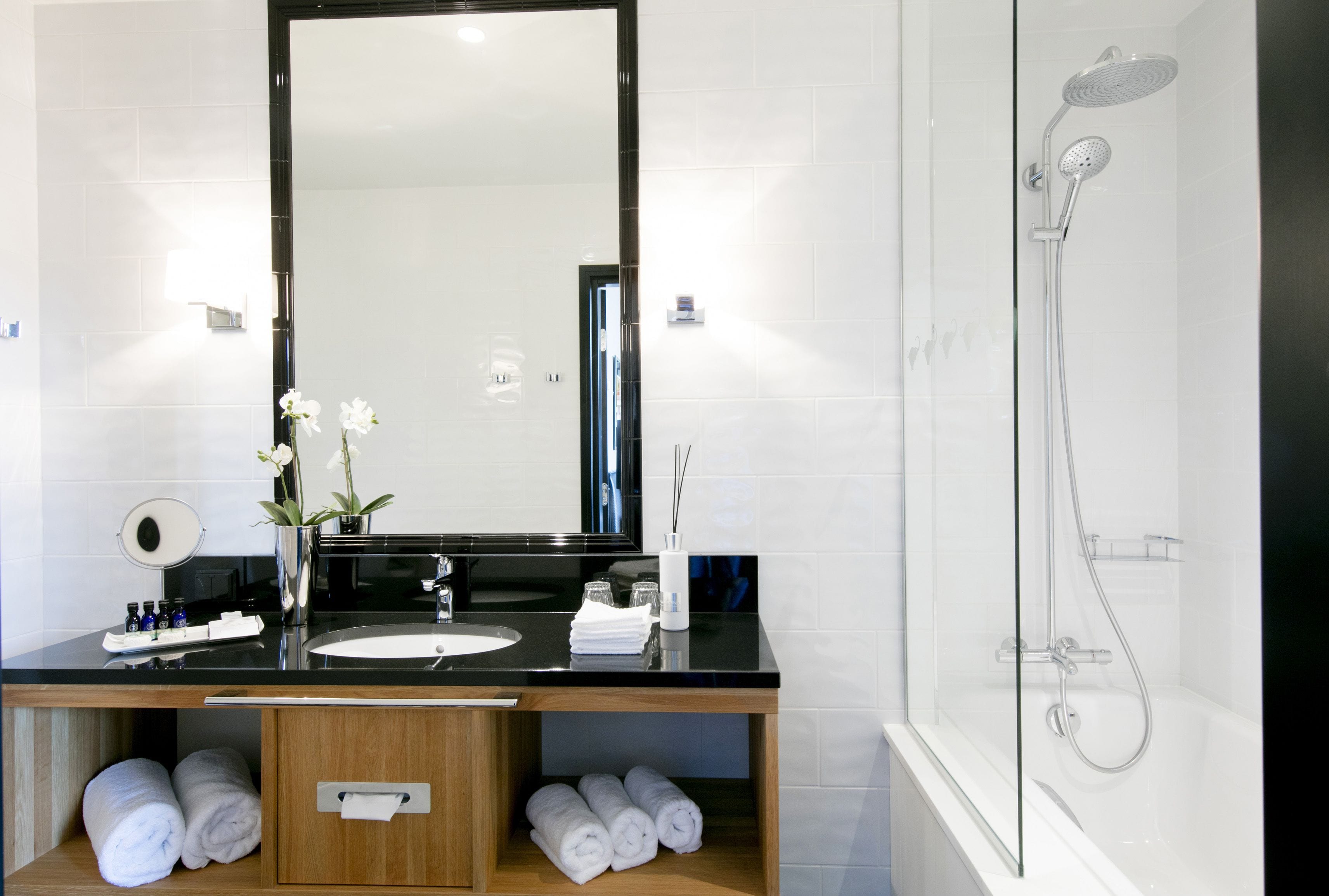 Une salle de bain moderne avec un lavabo à vasque sur un comptoir noir, un grand miroir, une douche à parois de verre et des serviettes soigneusement pliées.
