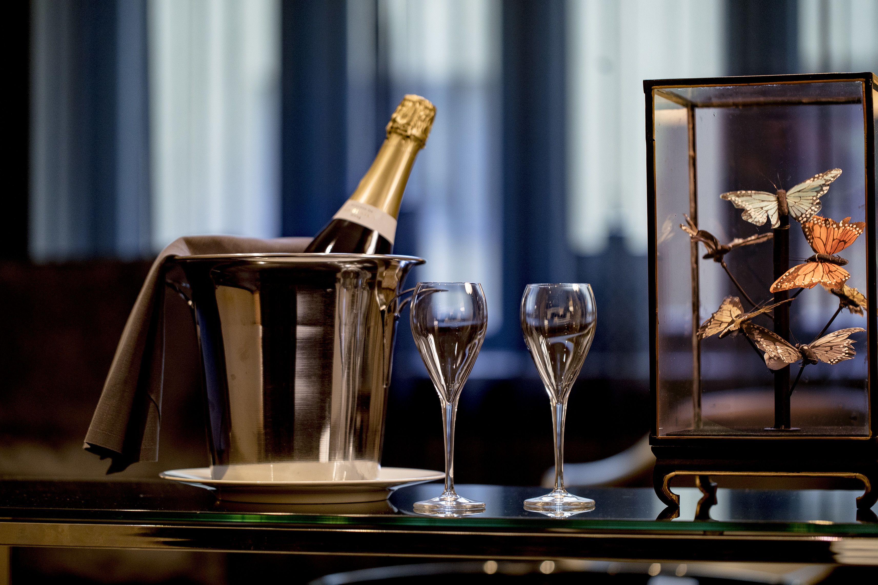 A champagne bottle in a silver bucket, two empty wine glasses, and a framed butterfly display.