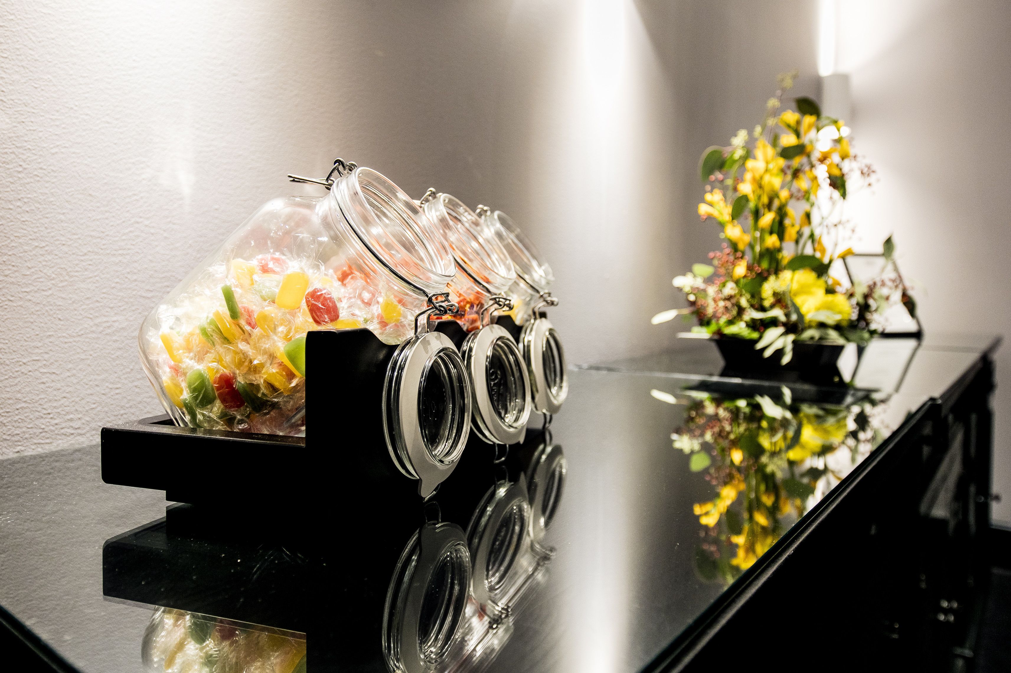 A buffet setup with a glass container holding assorted snacks and a flower arrangement on a reflective black table.