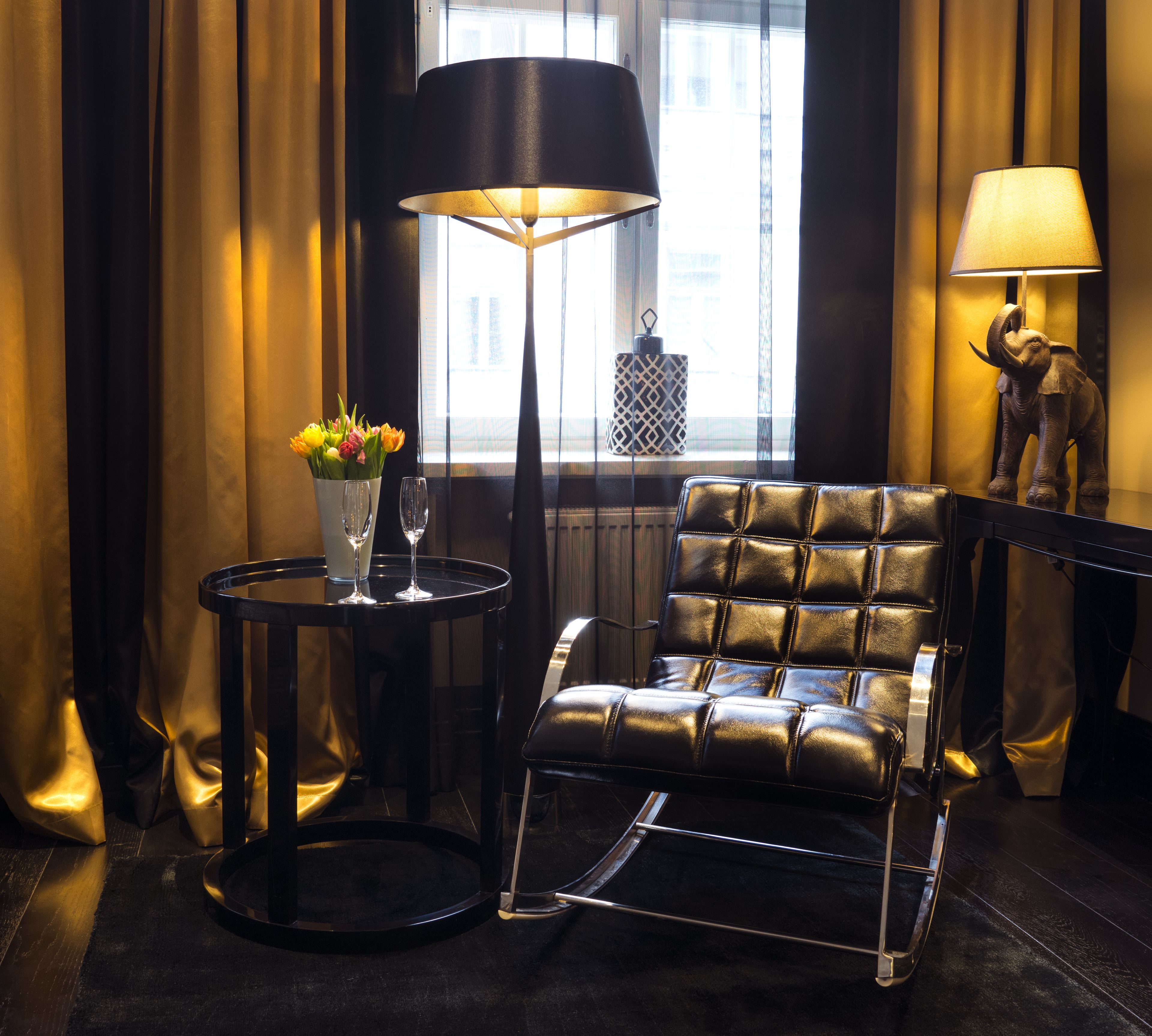 A leather armchair with a metal frame, a small round table with a vase of flowers and two wine glasses, a floor lamp, and a decorative elephant statue.