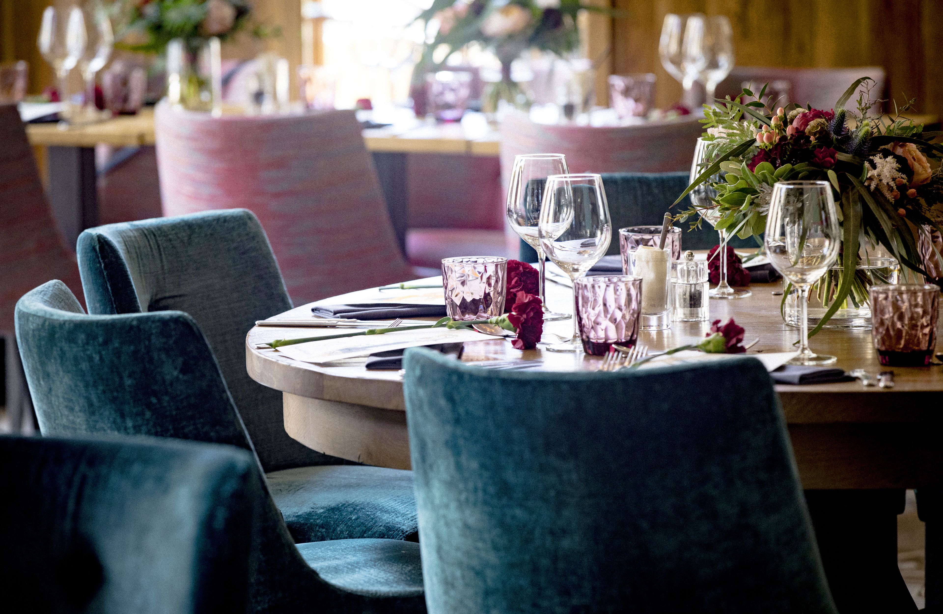Un comedor con mesas redondas dispuestas para dos personas, con sillas tapizadas en azul oscuro y un centro de mesa floral.