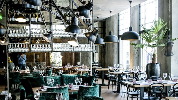 Interior de restaurante con mesas redondas, sillas de terciopelo verde e iluminación colgante.