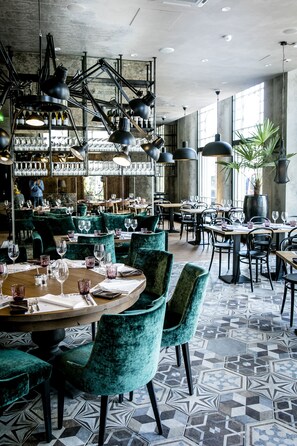 Interior de restaurante con mesas redondas, sillas de terciopelo verde e iluminación colgante.