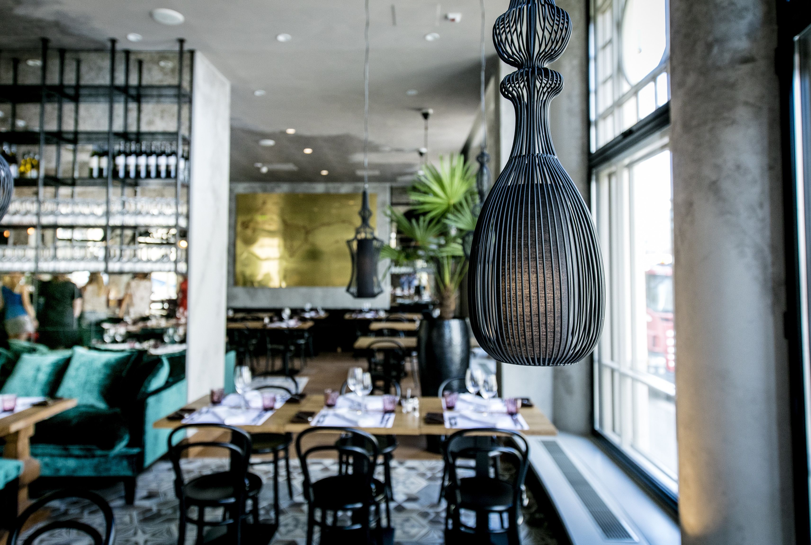 Un restaurante con mesas y sillas, una zona de bar con estantes llenos de botellas y lámparas colgantes.