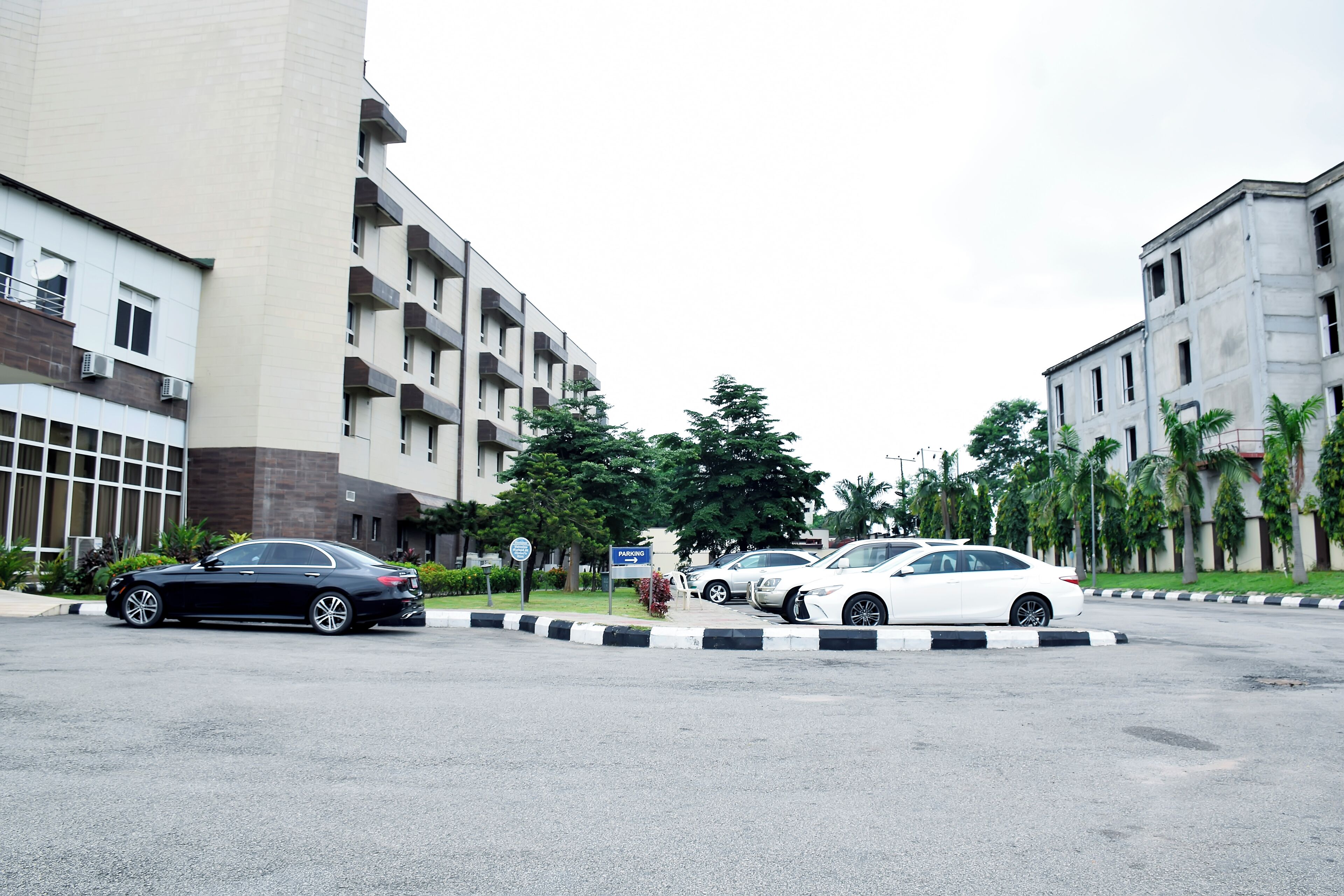 A parking area with cars, a building with multiple floors and balconies, and a sign indicating parking availability.