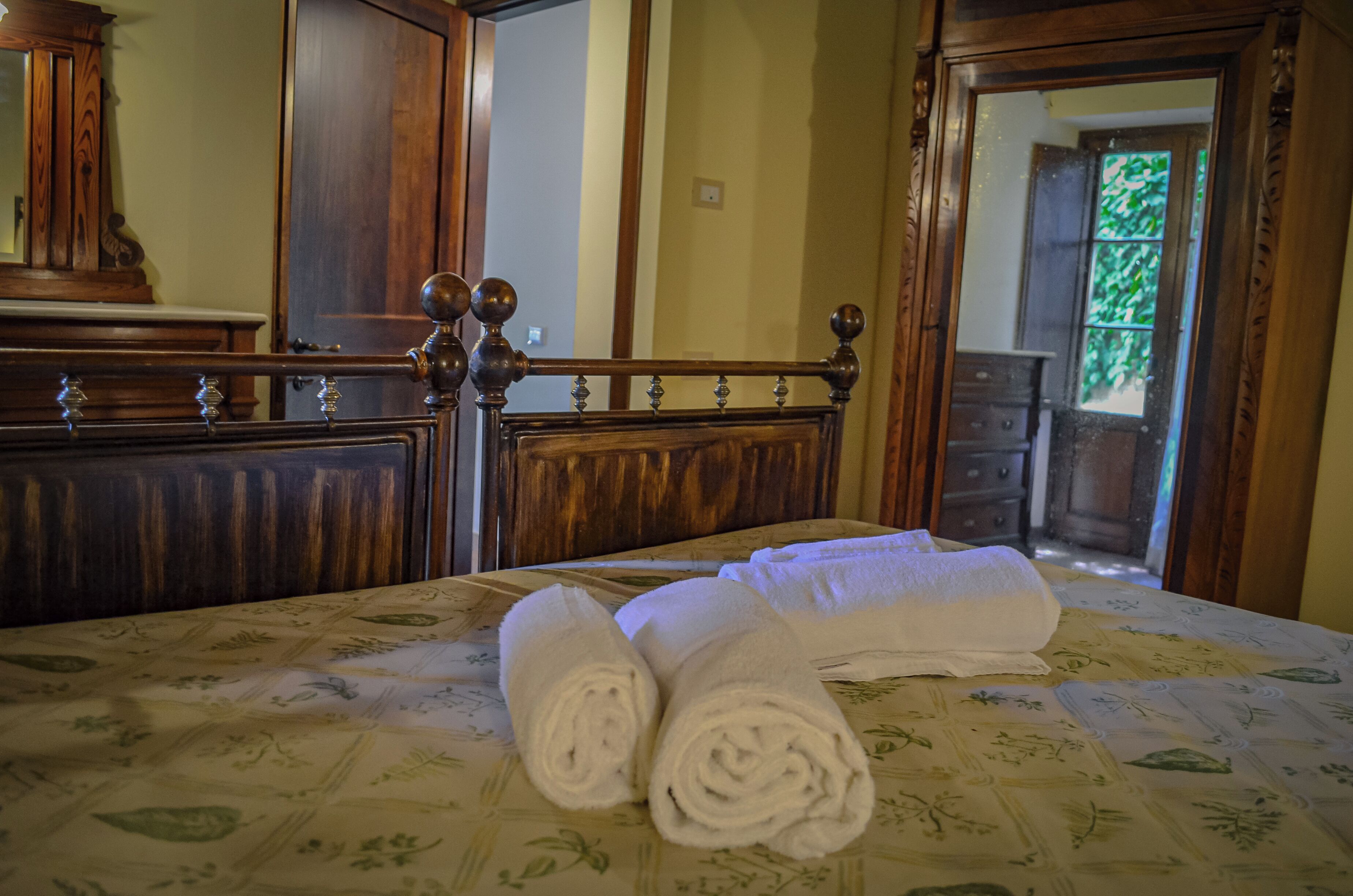 Un lit bien fait, avec des serviettes blanches pliées en rouleaux, une tête de lit en bois et une couvre-lit à motifs floraux.