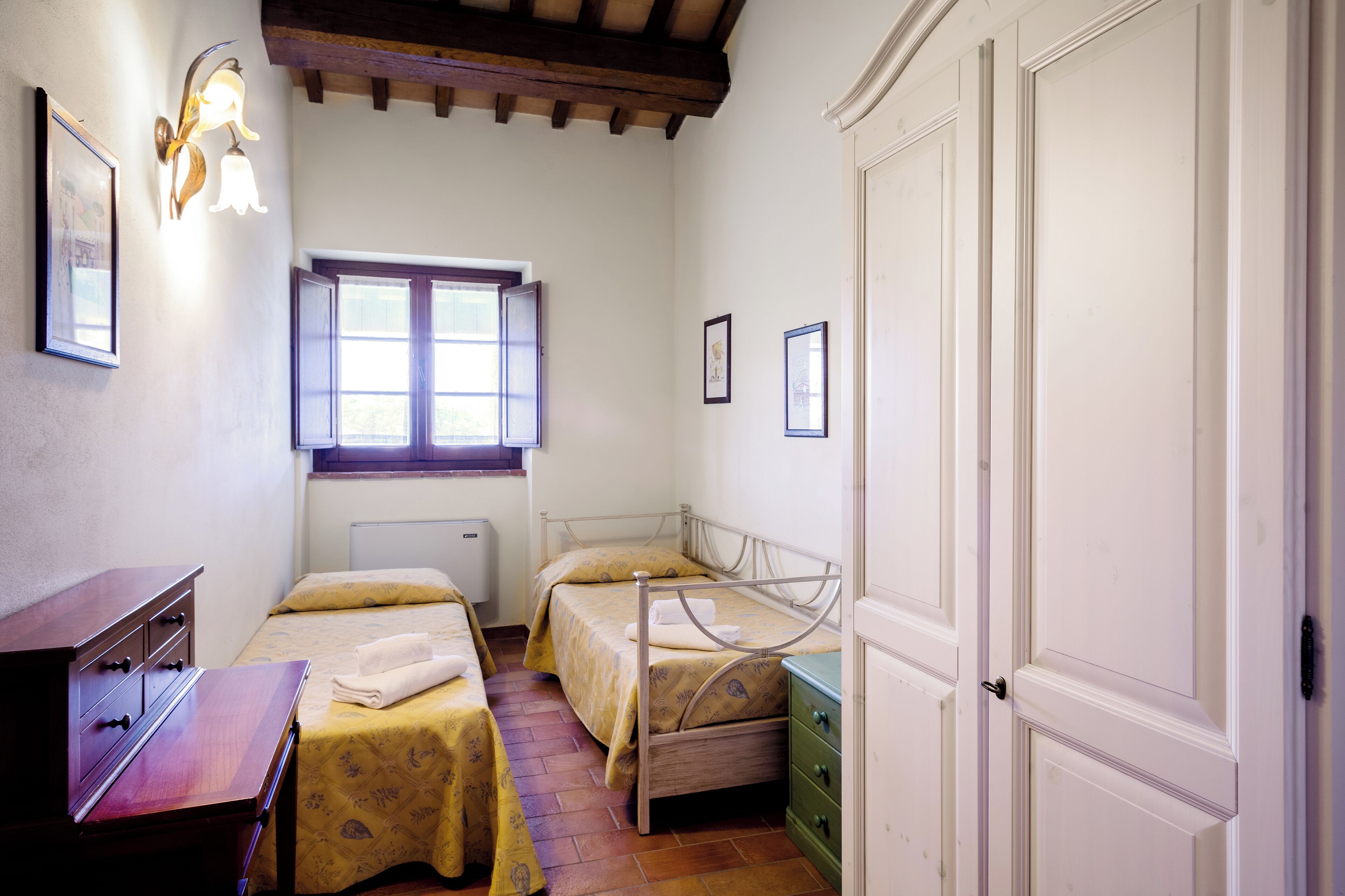 Quarto com duas camas de solteiro, uma cômoda de madeira, mesa de cabeceira, janela com persianas e porta.