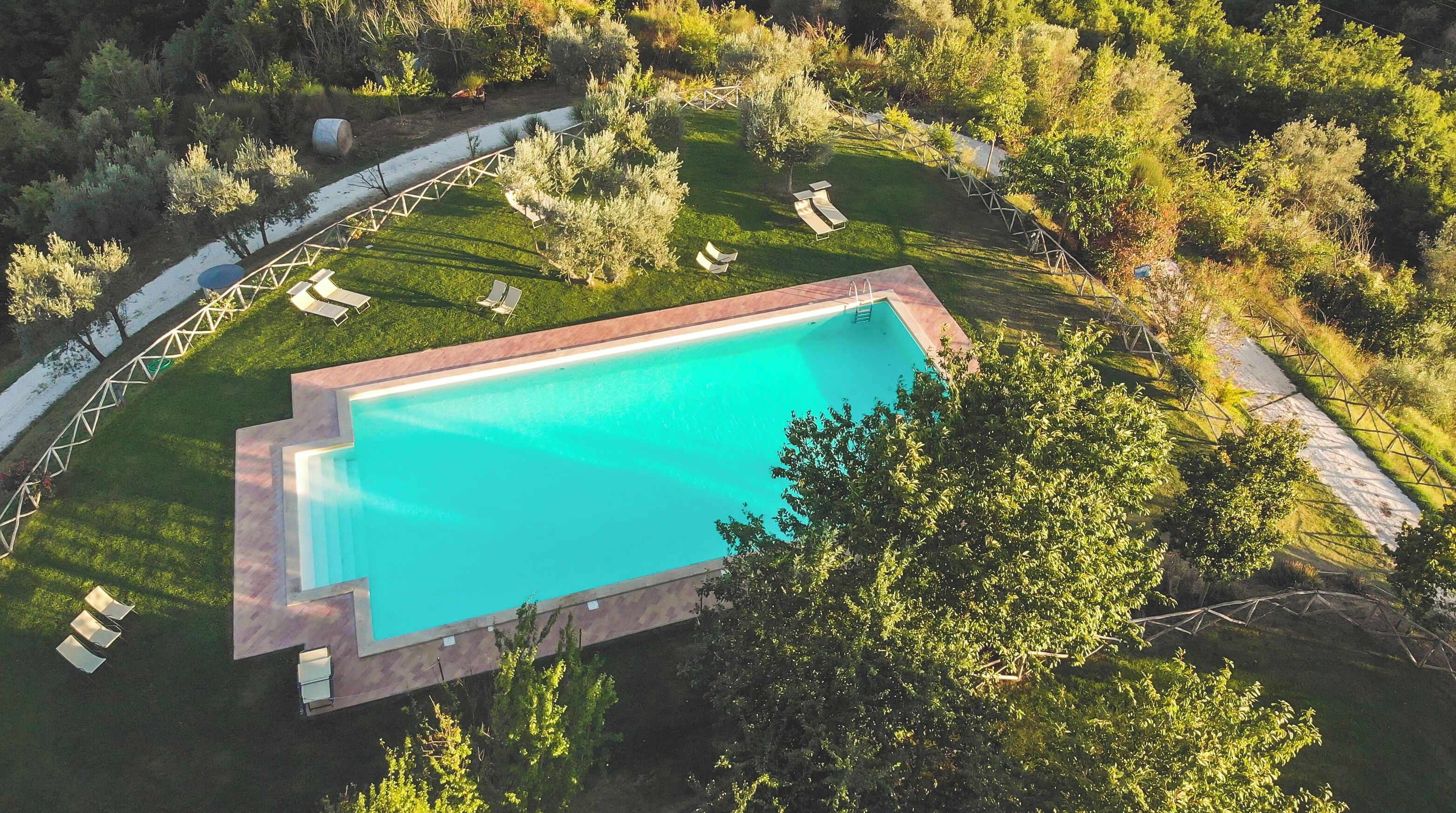 Vista aérea de una piscina rectangular rodeada por un césped con varias sillas de descanso y una terraza de madera.
