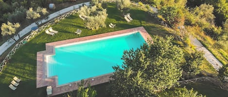 Vista aérea de una piscina rectangular rodeada por un césped con varias sillas de descanso y una terraza de madera.