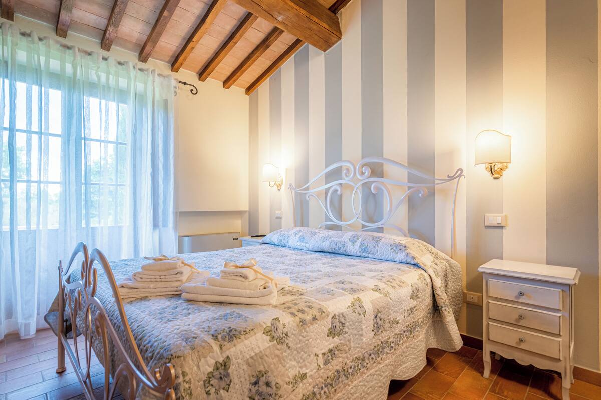Ein Schlafzimmer mit einem Metallbettgestell, einer blumigen Tagesdecke, einem Nachttisch mit Schubladen und einer Wandleuchte.