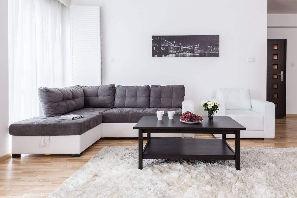 Un salon moderne avec un canapé d’angle gris, un canapé blanc, une table basse noire et une œuvre d’art représentant un paysage urbain accrochée au mur.