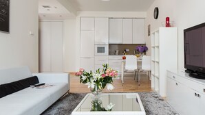 A modern living room with a sofa, a glass coffee table with flowers, a TV, and a kitchen in the background. - Chopin Apartments Platinum Towers (Warsaw)