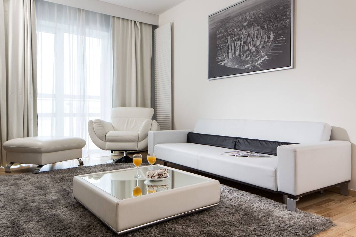 Un salon moderne avec un canapé blanc, une table basse en verre sur laquelle sont posées des boissons, un tapis gris et un grand tableau représentant un paysage urbain en noir et blanc accroché au mur.