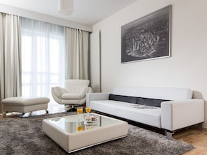 A modern living room with a white sofa, a glass coffee table with drinks, a grey rug, and a large black-and-white cityscape painting on the wall.