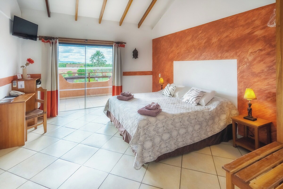 Un dormitorio con cama, lámparas de noche, escritorio, silla, televisor y vistas al exterior.