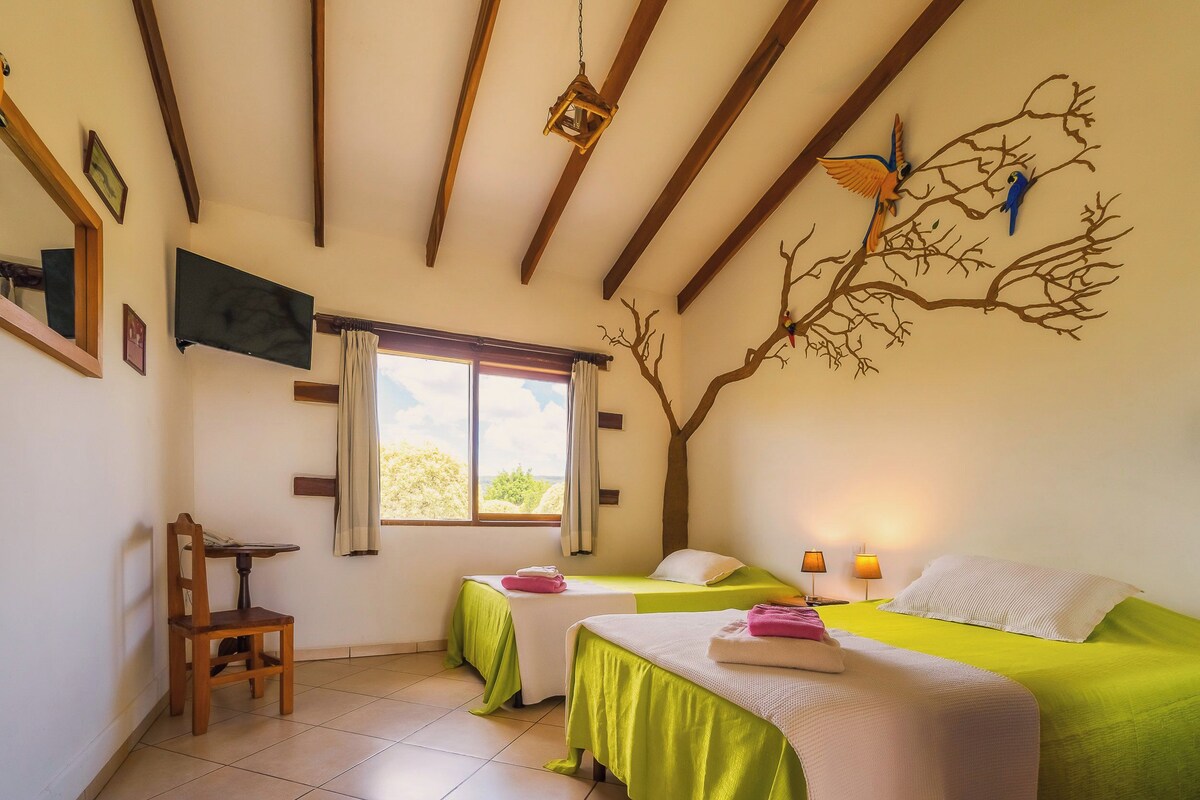 Una habitación con dos camas, una silla, una mesa, un ventanal y un mural en la pared.