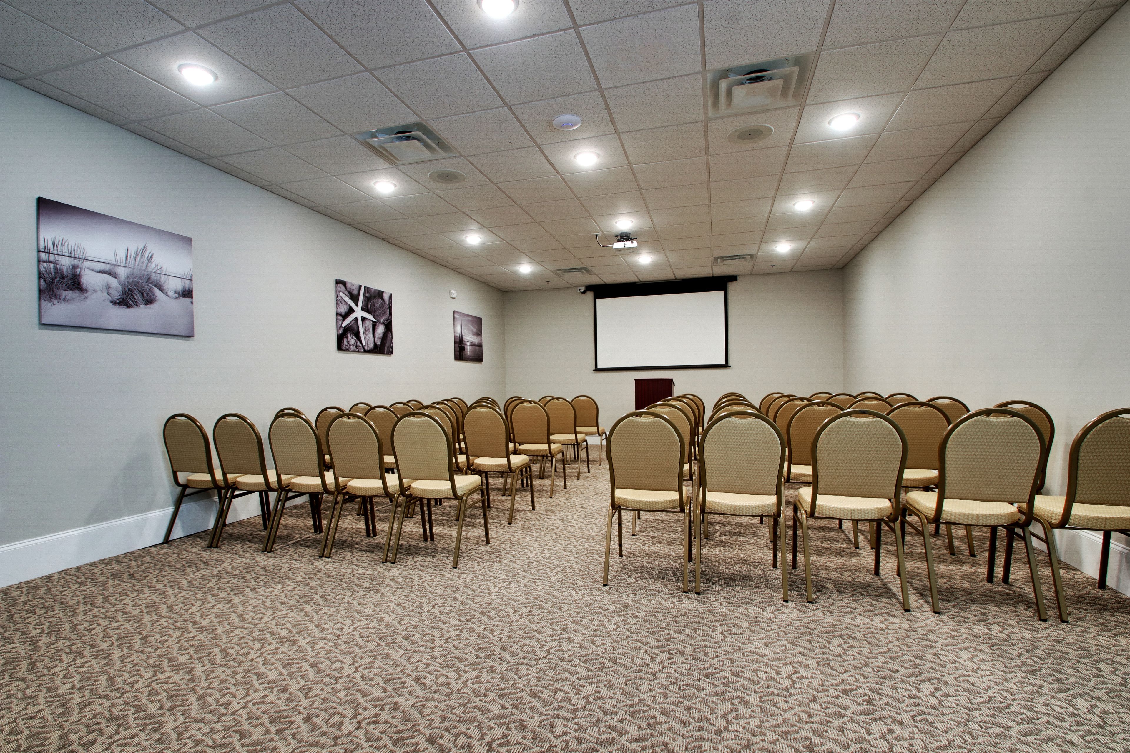 A conference room with rows of beige chairs, a carpeted floor, a projector screen, and artwork on the walls.